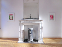 In the middle of the pictures is an old fireplace with a mirror on top, on the fireplace is a sculpture, right end left of it are two framed red and green watercolorpainting. Anna Slobodnik, 2018. 