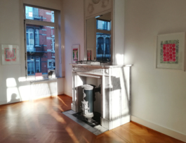 View of a room with a fireplace and a window on the left inbetween of them and on each side, three framed watercolor paintings in red and green. Anna Slobodnik, 2018. 