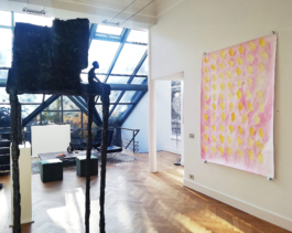 To the left a big metallsculpture of a man sitting in front of a house, on the right a big watercolorpainting of yellow shapes on pink ground. In the background a big wall of windows. Anna Slobodnik, 2018. 