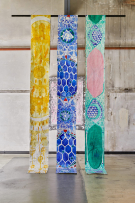 Three canvases hanging down in blue, yellow and green, behind them a work on paper. 