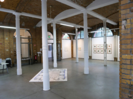 View of a room with columns, on the right framed painting are hanging on the wall. On the floor an assembled drawing is lying. 