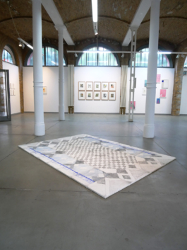 Exhibition view of a room with a big canvas drawing lying on the floor, behind it on the wall are framed paintings. 