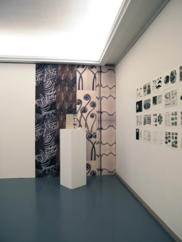 Drawings on a wall to the right, to the left, a wallpaper covered wall with a plinth in front of it and a book on the plinth. 