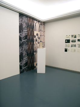 Drawings on a wall to the right, to the left, a wallpaper covered wall with a plinth in front of it and a book on the plinth. 