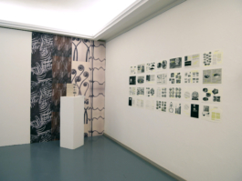 Drawings on a wall to the right, to the left, a wallpaper covered wall with a plinth in front of it and a book on the plinth. 