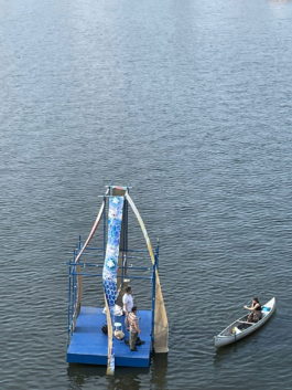 Two people on a raft and the Artist on a canoe. On the raft: one ornamental blue pattern. 