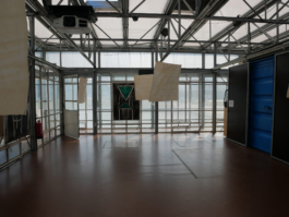 View of an exhibition with an oil painting of a chair in the back and paper works hanging from the ceiling in the front. 