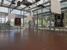 View of an exhibition with drawings on paper hanging from the ceiling with three oil paintings of chairs hanging on the glass walls. On the outside are trees and buildings. 