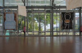 Installation view of two chair paintings a blue striped one and a beige one and paper works hanging from the ceiling. 