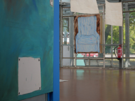 In the front left corner a detail of a painting of a turquoise door to the right of it there is a room opening up in it drawings on paper hanging from the ceiling and a painting of a blue striped arm chair. 