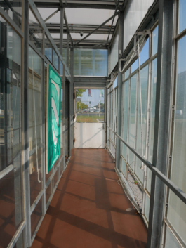 A passage in a glassbuilding at the end of it a door. On the left wall there is a painting of a green glass. 