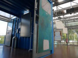 Frog perspective on the entrance of an exhibition space with a turquoise door in the middle, the entrance to the left and the space to the right. 