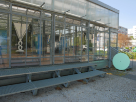 View of a building made out of glass with paintings of glasses in it. To the right there is an entrance with some stairs and a green round table leaning on them. 