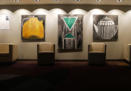 For armchairs standing in front of a wall with three paintings of two teapots and a chair in the middle. 