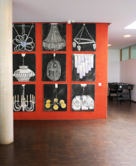 Front view of 9 paintings of lamps mostly in black and white hanging on a red wall. 